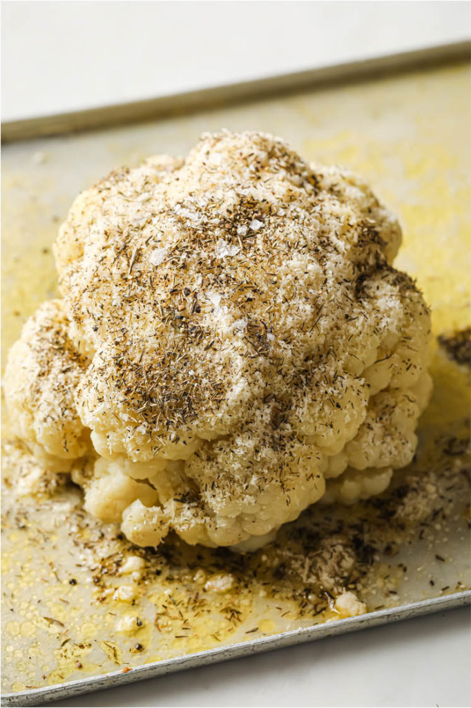 Side shot of cauliflower head drizzled with melted butter, grated Parmesan, thyme, salt, and pepper on a baking pan