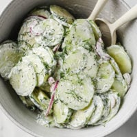 Overhead shot of Creamy Dill Cucumber Salad in a ceramic bowl with two mixing spoons atop a marble countertop