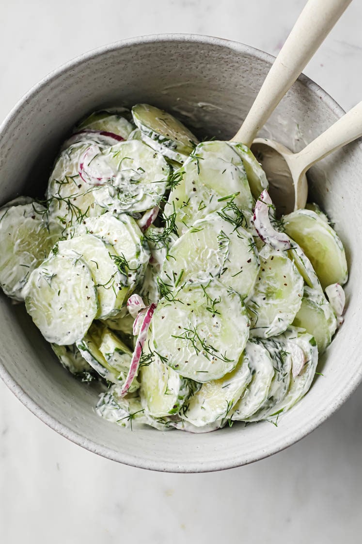 Overhead shot of Creamy Dill Cucumber Salad in a ceramic bowl with two mixing spoons atop a marble countertop