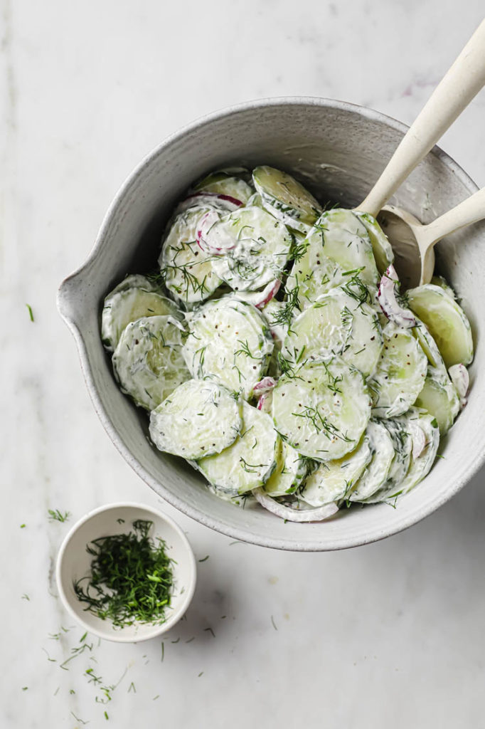 Overhead shot of cucumber salad in a ceramic bowl with two mixing spoons and a small bowl with fresh dill for additional garnish
