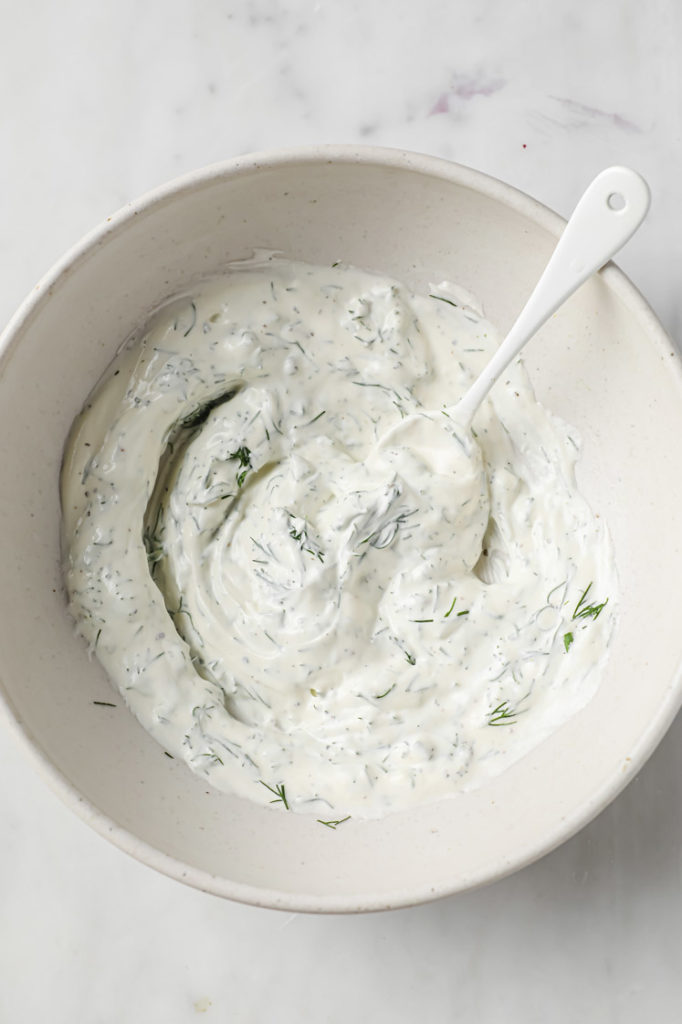Overhead shot of mixed sour cream, mayonnaise, dill, and pepper with a white spoon in a ceramic bowl
