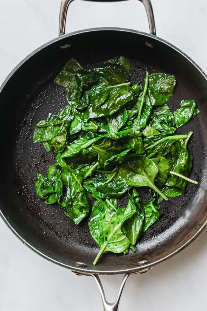Overhead shot of fresh spinach being cooked on a frying pan. The frying pan is resting on a marble countertop.