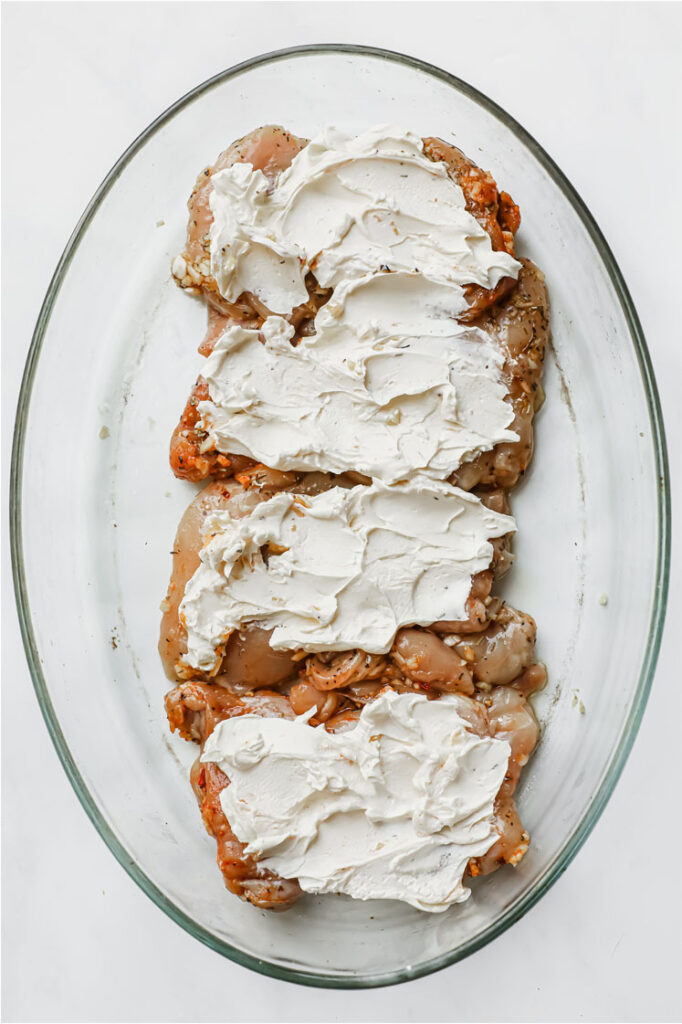 Overhead shot of marinated chicken thighs with softened cream cheese spread atop in a glass baking pan. The baking pan is resting on a marble countertop.