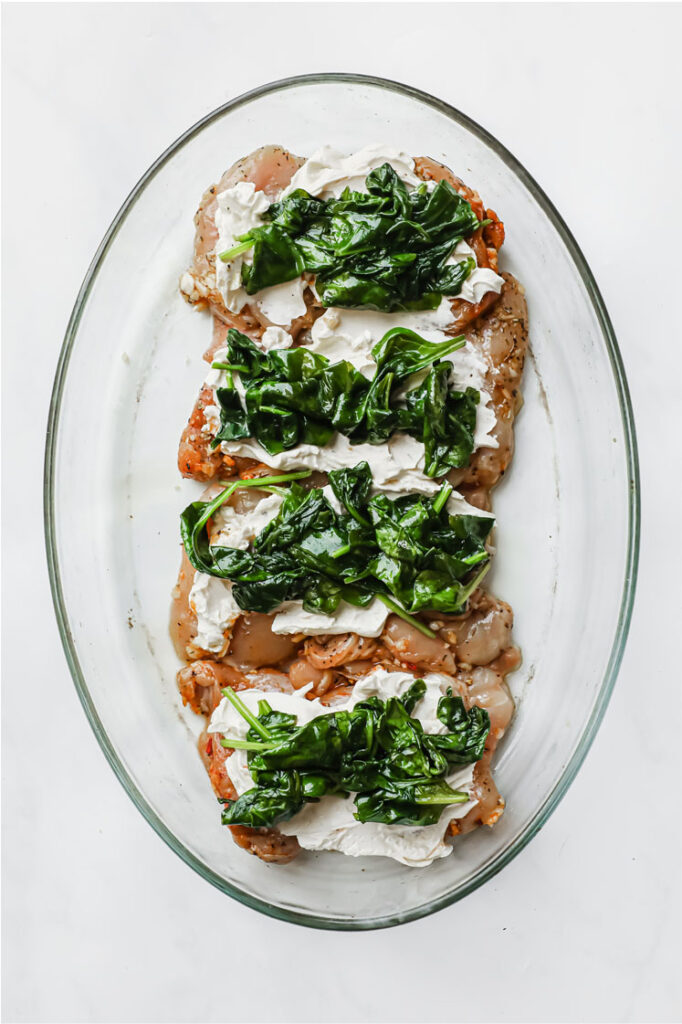 Overhead shot of marinated chicken thighs with softened cream cheese and cooked spinach spread atop in a glass baking pan. The baking pan is resting on a marble countertop.