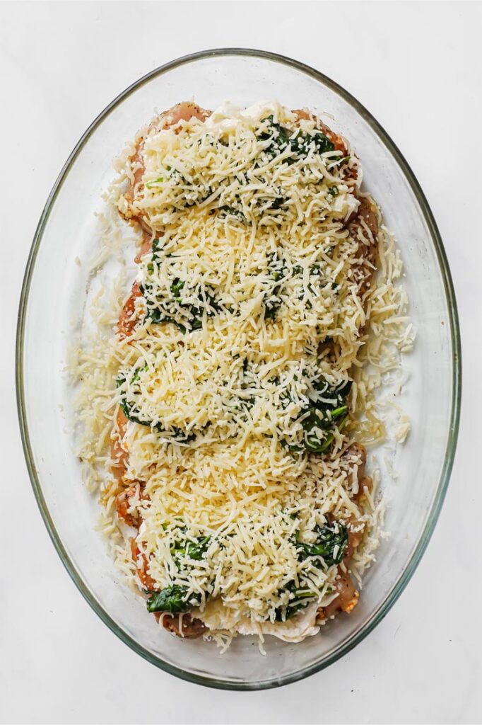 Overhead shot of marinated chicken thighs with softened cream cheese, cooked spinach and mozzarella spread and sprinkled atop in a glass baking pan. The baking pan is resting on a marble countertop.