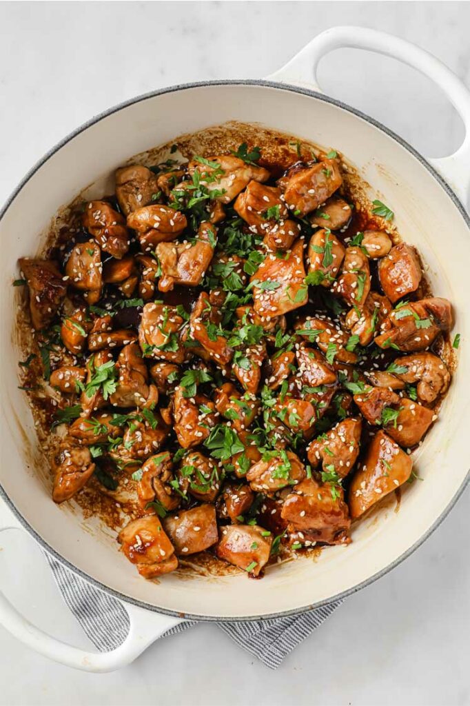 Overhead shot of a large pan with chicken thighs, garnished with freshly-cracked pepper, sesame seeds, and fresh parsley. The large pan is resting on a marble countertop.