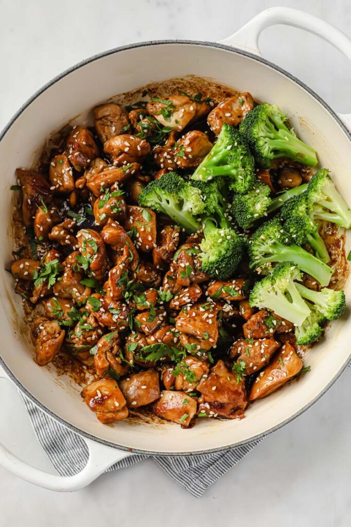 Overhead shot of a large pan with the sweet sticky chicken thighs paired with broccoli and garnished with freshly-cracked pepper, sesame seeds, and fresh parsley.
