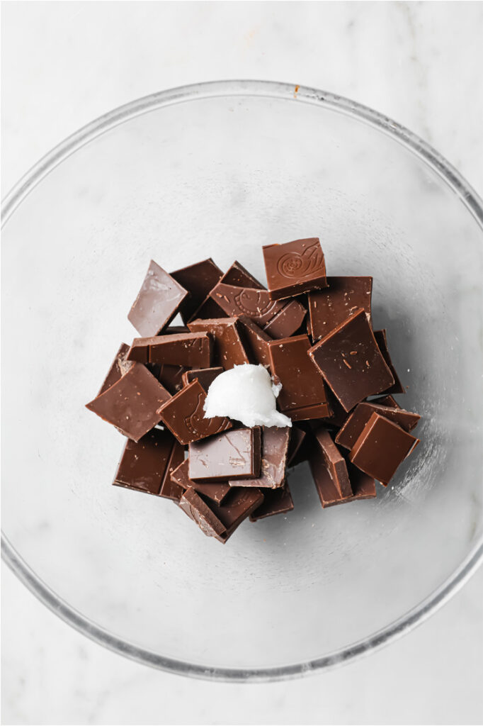 Overhead shot of chocolate bar broken into small pieces and coconut oil in a microwave-safe bowl, atop a marble counter.