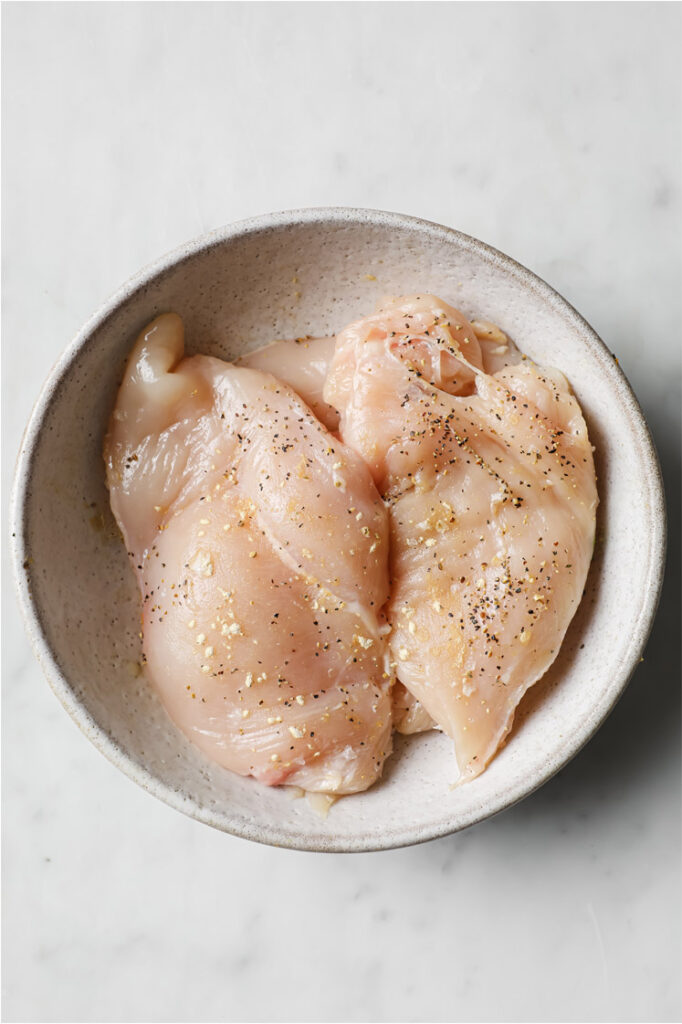 Overhead shot of a small bowl with chicken breasts, seasoned with garlic powder, pepper, and salt. The small bowl sits atop a marble counter.