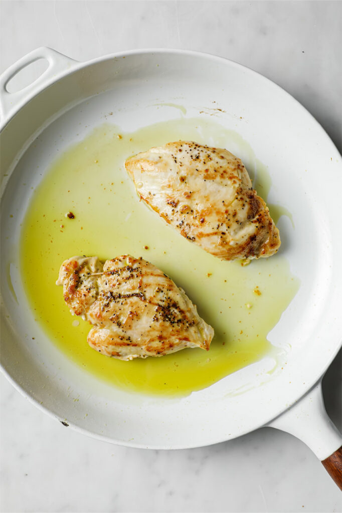 Overhead shot of a pan with chicken breasts, cooked in oil until through and golden. The pan sits atop a marble counter.