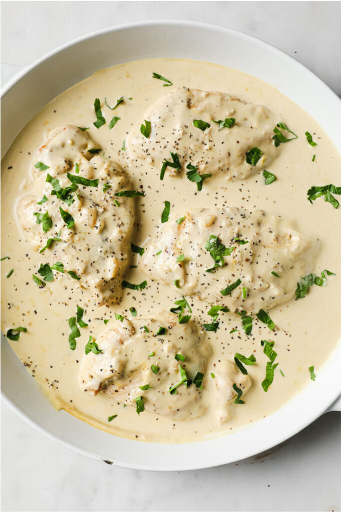 Overhead shot of a pan with creamy garlic chicken, garnished with fresh parsley and freshly-cracked black pepper. The pan sits atop a marble counter.
