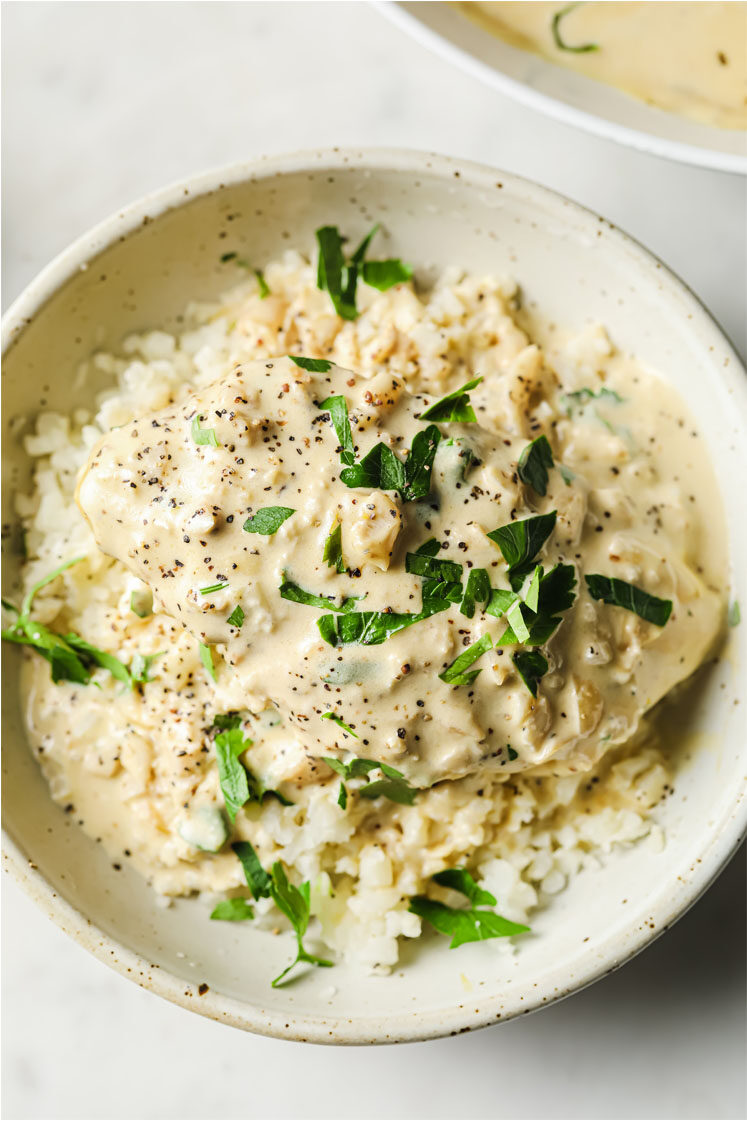 Overhead shot of a small bowl with creamy garlic chicken served on top of cauliflower rice, garnished with fresh parsley and freshly-cracked black pepper. The small bowl sits atop a marble counter.