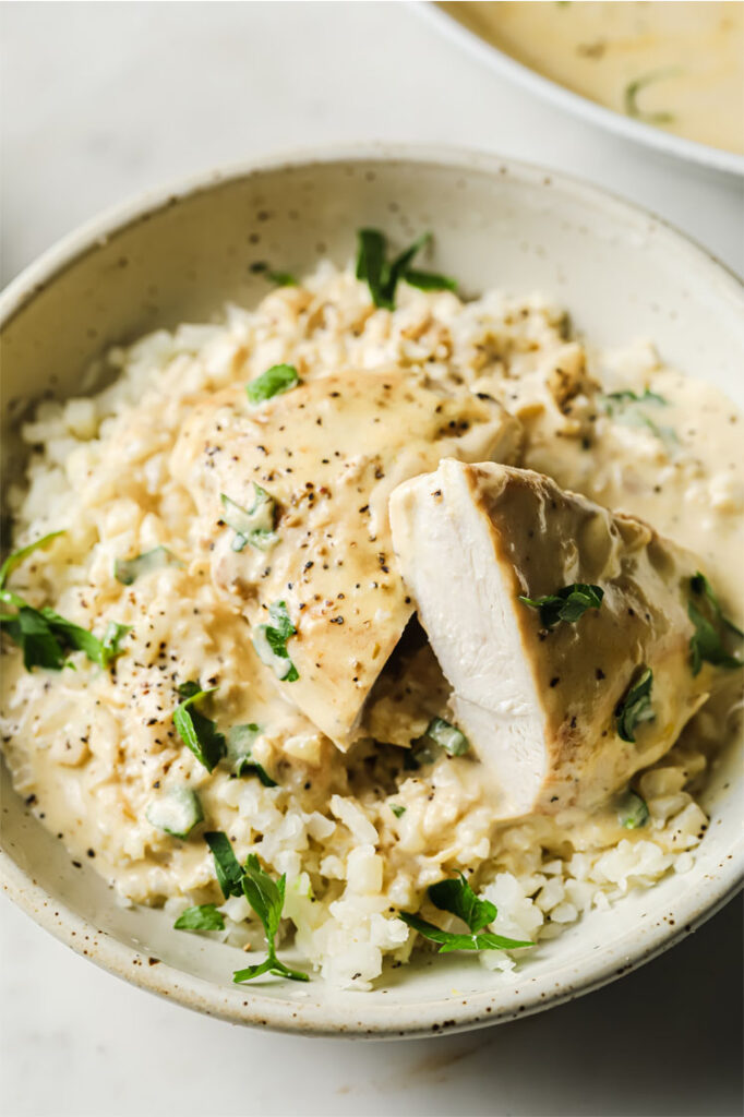 Overhead shot of a small bowl with creamy garlic chicken served on top of cauliflower rice, garnished with fresh parsley and freshly-cracked black pepper. The small bowl sits atop a marble counter.