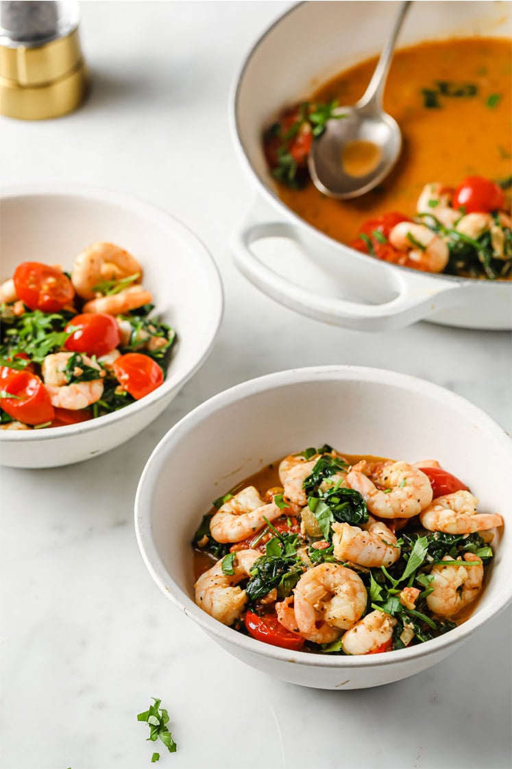 Overhead shot of the One-Pan Low-Carb Tuscan Garlic Shrimp, garnished with chopped fresh parsley and Italian seasoning, served in small bowls, atop a marble kitchen counter. In the background, you can see a cooking pan with the remaining One-Pan Low-Carb Tuscan Garlic Shrimp and a serving spoon.