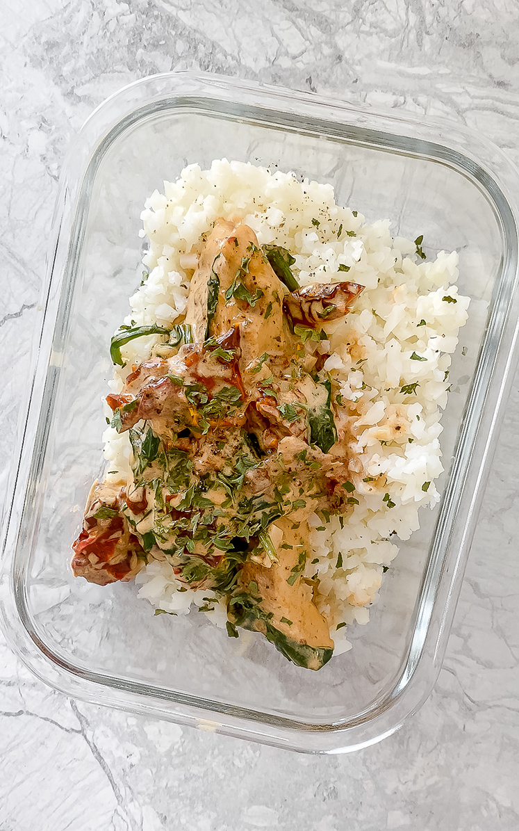 overhead shot of meal prep container with tuscan chicken and cauliflower rice atop marble countertop