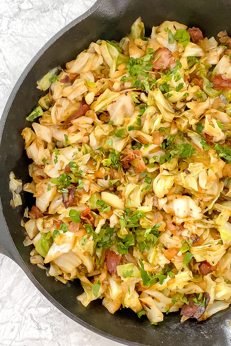 overhead shot of sauteed cabbage with bacon and parsley in cast iron skillet atop marble countertop