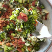 Close up shot of a bowl and a fork with low-carb cheesy broccoli and bacon, atop a marble countertop.