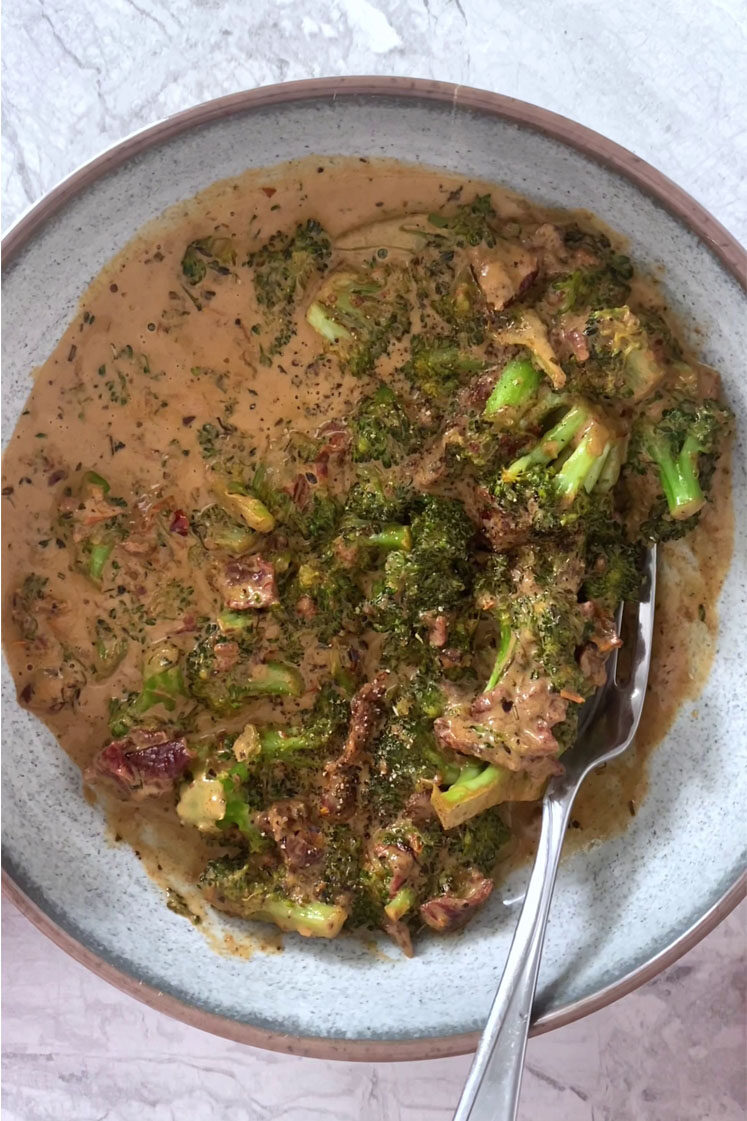 Overhead shot of a ceramic bowl filled with creamy italian broccoli garnished with freshly-cracked black pepper. The ceramic bowl has a fork in it and it sits atop a marble countertop.