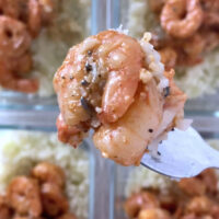 Overhead shot of a fork with a portion of the meal prep cajun shrimp and cauliflower rice hovering over four glass meal-prep containers with the meal prep cajun shrimp and cauliflower rice, drizzled with sauce. The glass containers sit atop a marble countertop.