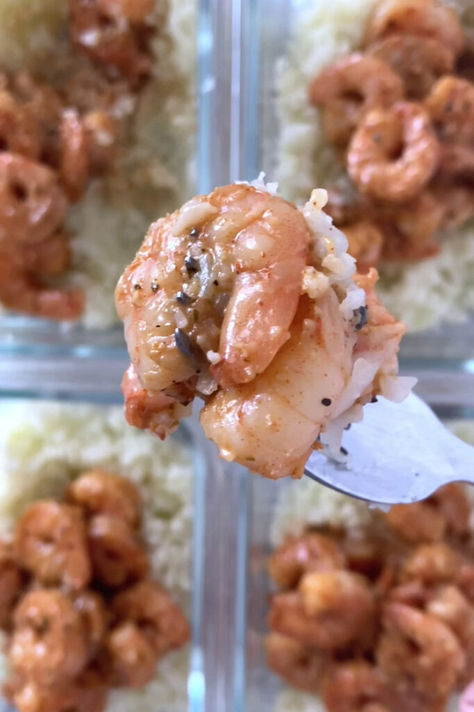 Overhead shot of a fork with a portion of the meal prep cajun shrimp and cauliflower rice hovering over four glass meal-prep containers with the meal prep cajun shrimp and cauliflower rice, drizzled with sauce. The glass containers sit atop a marble countertop.