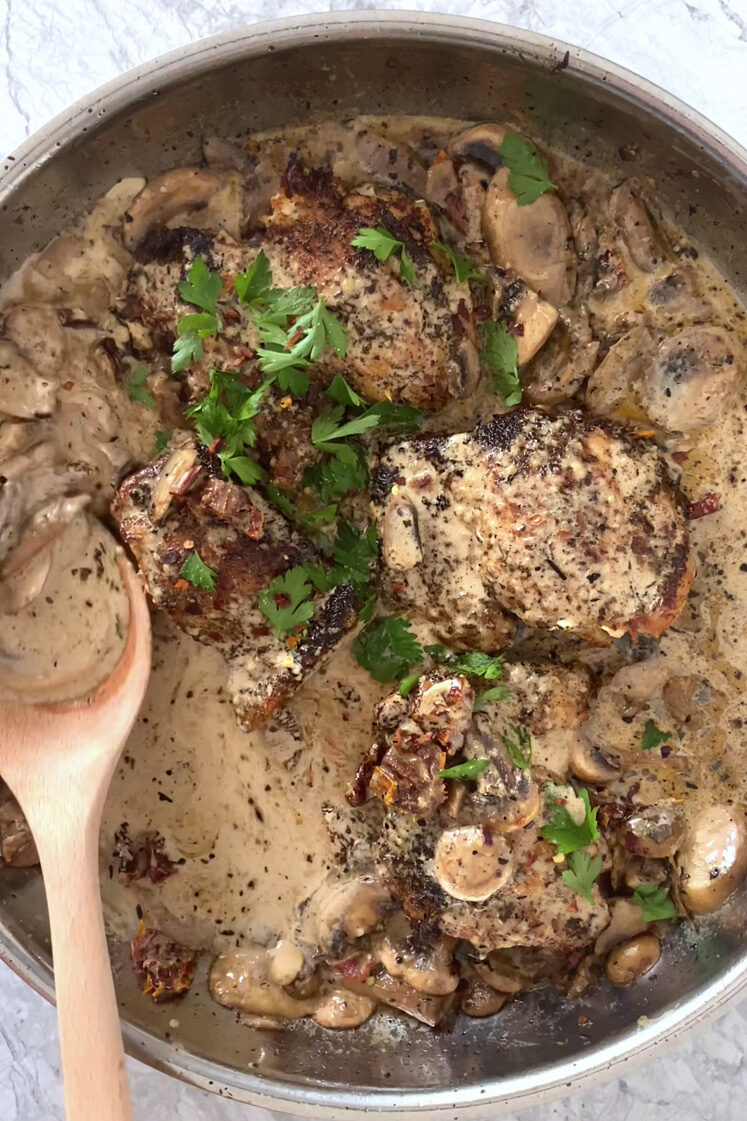 Overhead shot of a pan with the creamy mushroom sauce chicken thighs garnished with freshly-cracked pepper, red pepper flakes, and fresh parsley. The pan has a wooden serving spoon and sits atop a marble countertop.