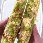 Overhead, close-up shot two cucumber boats filled with egg salad and garnished with green onions, paprika, cayenne pepper, flakey sea salt, and cracked pepper. A cutting board can be seen in the background, and it rests atop a marble countertop.