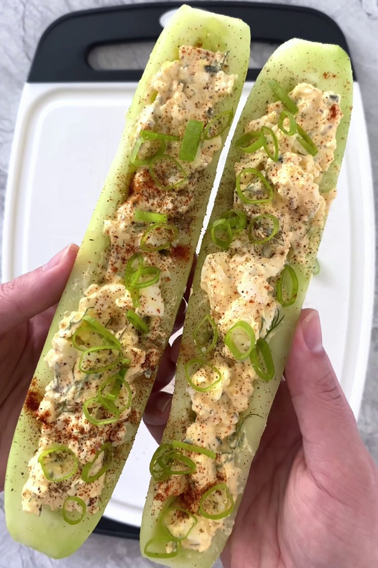 Overhead, close-up shot two cucumber boats filled with egg salad and garnished with green onions, paprika, cayenne pepper, flakey sea salt, and cracked pepper. A cutting board can be seen in the background, and it rests atop a marble countertop.