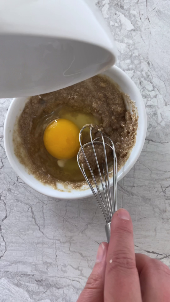 Overhead shot of a bowl with mixed dry ingredients and melted butter along with one raw egg, about to be mixed using a whisk. The bowl sits atop a marble countertop.
