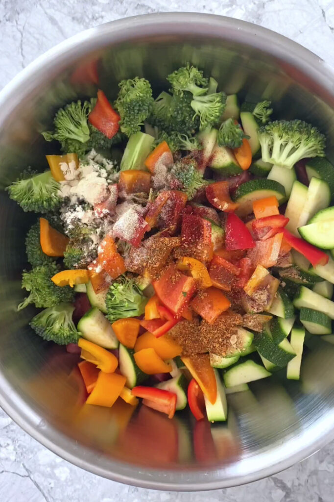 Overhead shot of a mixing bowl with sausage, vegetables, avocado oil, and spices. The bowl sits atop a marble countertop.