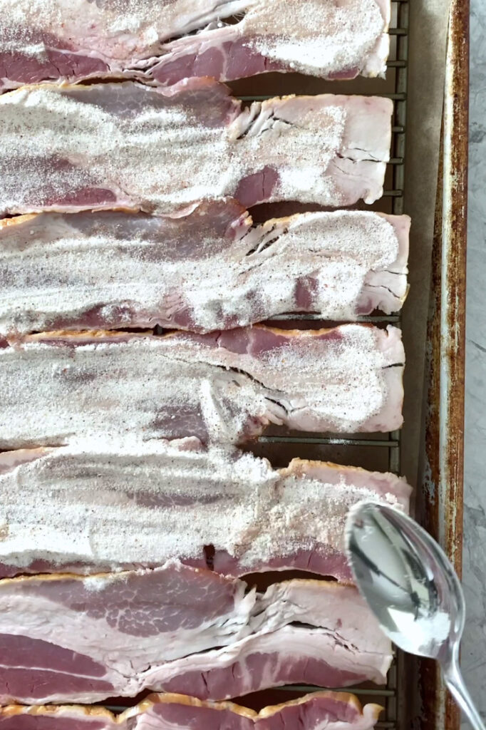 Overhead shot of bacon slices topped with allulose mixture using a spoon. The bacon slices are arranged in an even layer, and the wire rack rests inside a parchment paper-lined baking sheet.