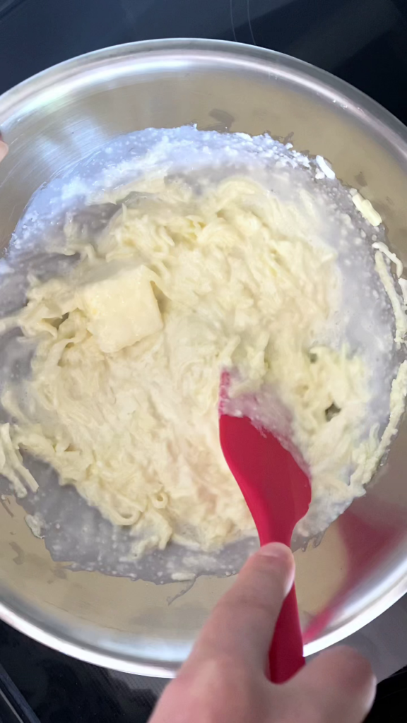 Overhead shot of a stainless steel bowl with shredded mozzarella, heavy cream, grated parmesan, and butter, set atop saucepan of simmering water, being stirred and melted together using spatula.