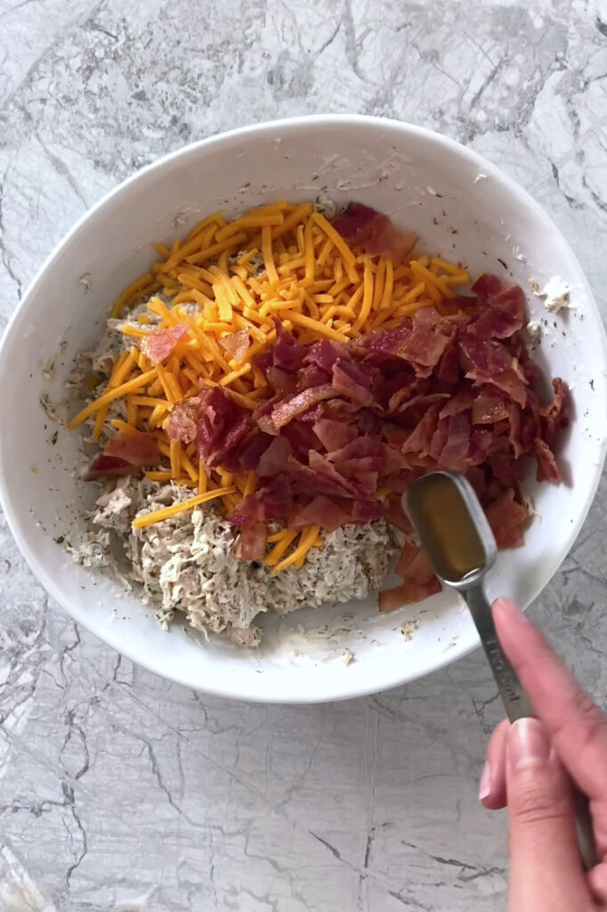 Overhead shot of a bowl with chicken and sour cream mixture, shredded cheddar, crumbled bacon, and apple cider vinegar. The bowl sits atop a marble countertop.