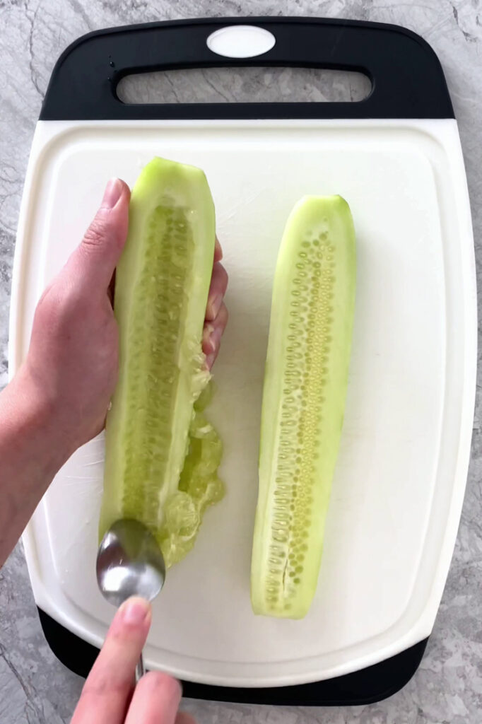 Overhead shot of a cutting board with one cucumber cut in half, lengthwise, atop a marble countertop. The seeds are removed using a spoon.