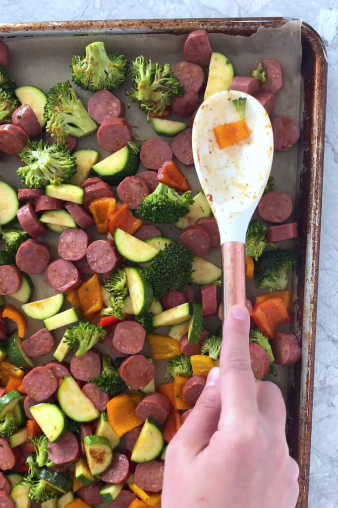 Overhead shot of a parchment paper-lined baking sheet with the sausage and vegetables spread to an even layer using a serving spoon. The pan sits atop a marble countertop.