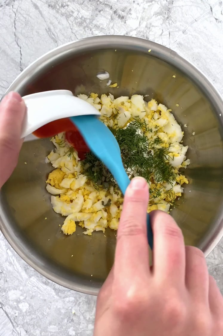 Overhead shot of a bowl with cooked and peeled eggs, Greek yogurt, dill, mustard, and chives. Hot sauce is added as well using a rubber spatula. The bowl sits atop a marble countertop.