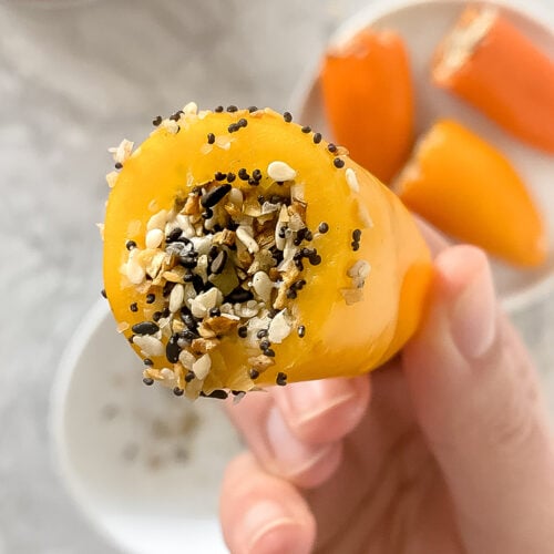 overhead shot of hand holding prepared 5-minute mini peppers topped with Everything Bagel Seasoning over top of plate of other prepared peppers atop marble countertop