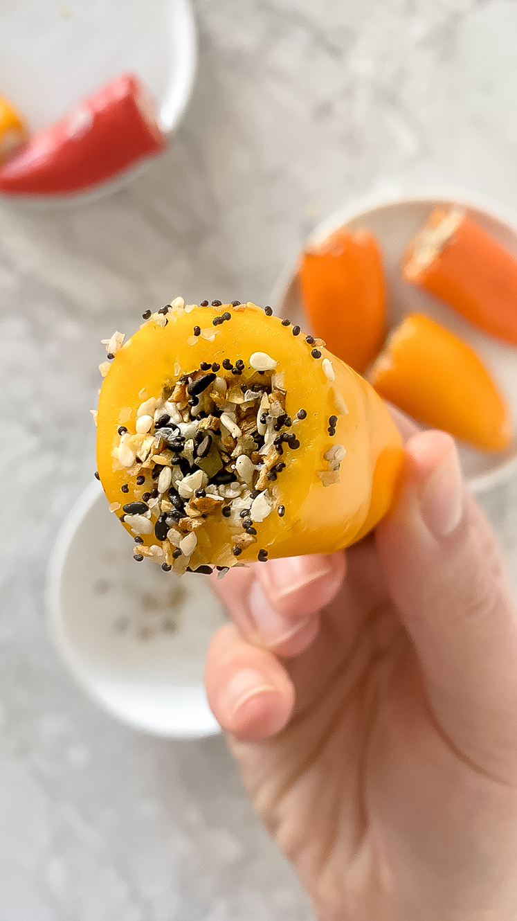 overhead shot of hand holding prepared 5-minute mini peppers topped with Everything Bagel Seasoning over top of plate of other prepared peppers atop marble countertop