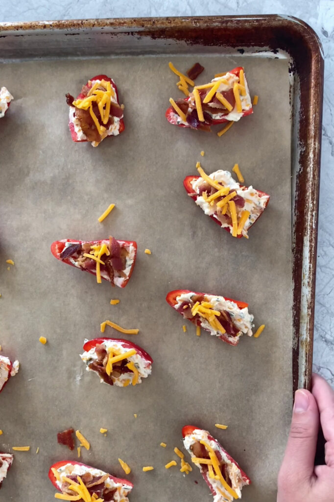 Overhead shot of a parchment paper-lined baking sheet with the halved mini-peppers filled with the cream cheese mixture, crumbled bacon, and remaining shredded cheddar.