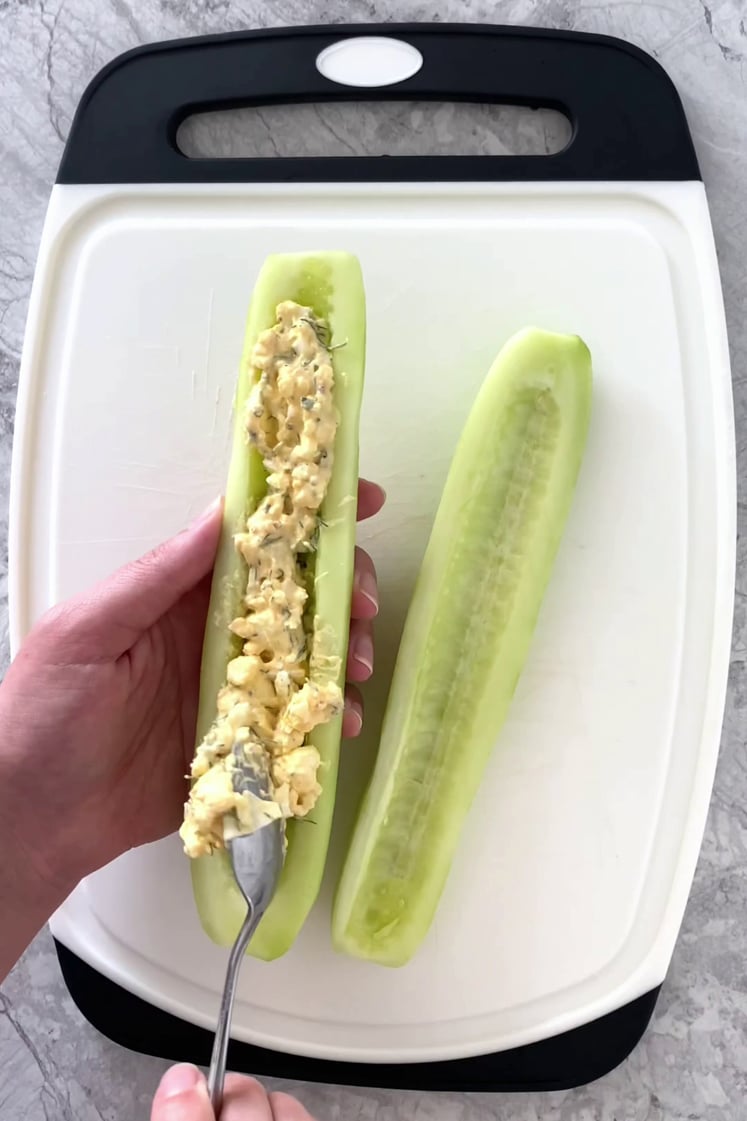 Overhead shot of a cutting board with two cucumber boats atop a marble countertop. The egg salad is spooned into one cucumber boat.