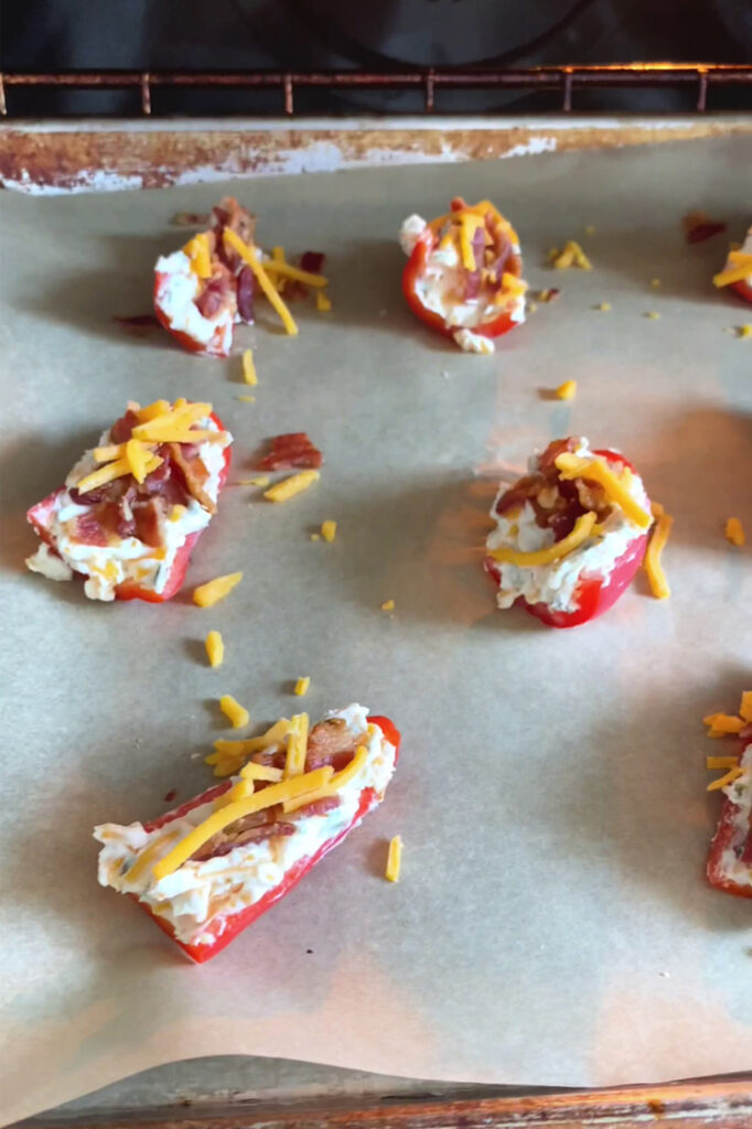 Angled shot of a parchment paper-lined baking sheet with the halved mini-peppers filled with the cream cheese mixture, crumbled bacon, and remaining shredded cheddar. The baking sheet is inside the oven.
