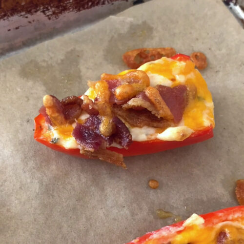 Close-up shot of the bacon and chive mini pepper poppers, fresh out from the oven. The bacon and chive mini pepper poppers rests inside a parchment paper-lined baking sheet. The baking sheet rests atop a marble countertop.
