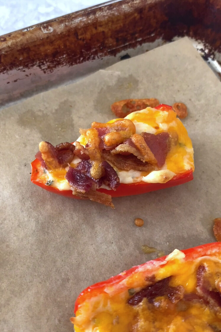 Close-up shot of the bacon and chive mini pepper poppers, fresh out from the oven. The bacon and chive mini pepper poppers rests inside a parchment paper-lined baking sheet. The baking sheet rests atop a marble countertop.