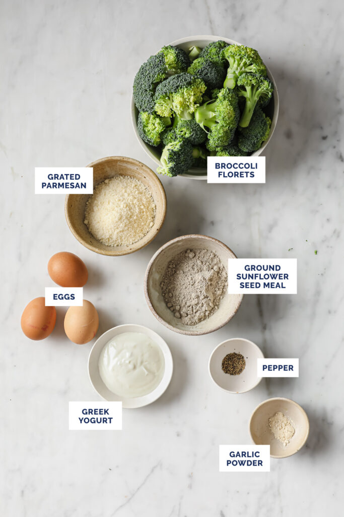 Overhead shot of labeled ingredients for the Broccoli Fritters, in bowls and plates, atop a marble countertop.