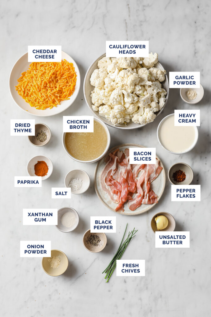 Overhead shot of labeled ingredients for the Cauliflower Cheddar Cheese Bacon Soup, in bowls and plates, atop a marble countertop.