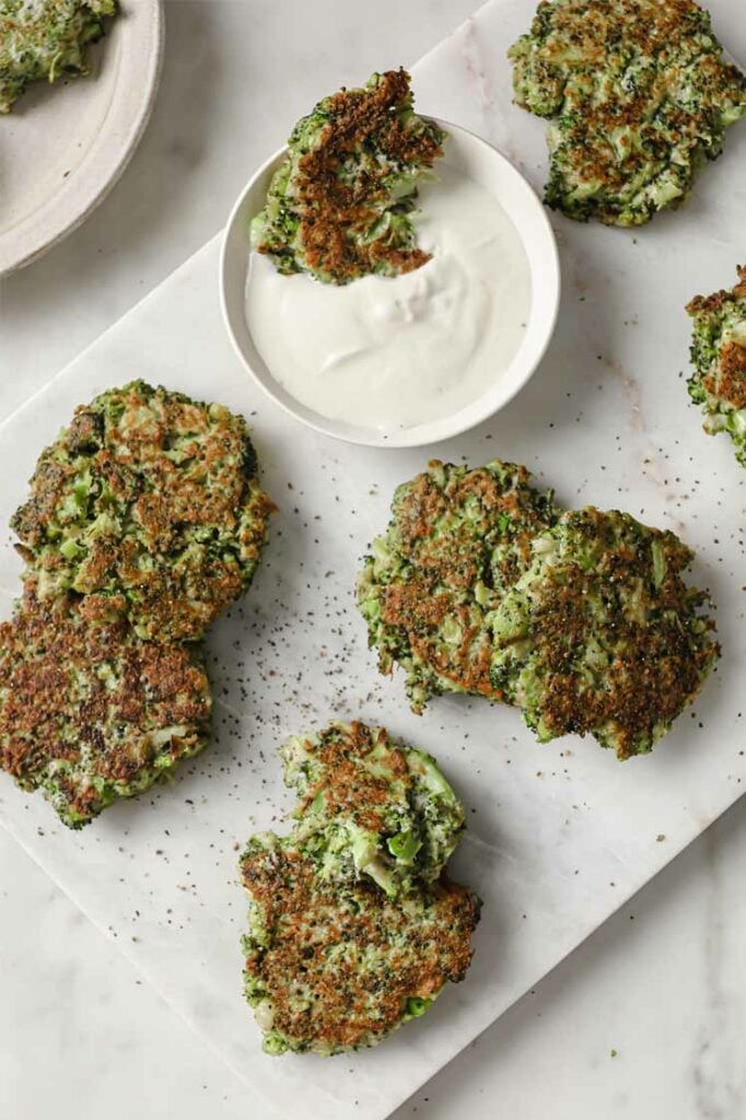 Overhead shot of a marbled plate with golden brown broccoli fritters and sour cream dip. One broccoli fritter is dipped in sour cream. The plate rests atop a marble countertop.
