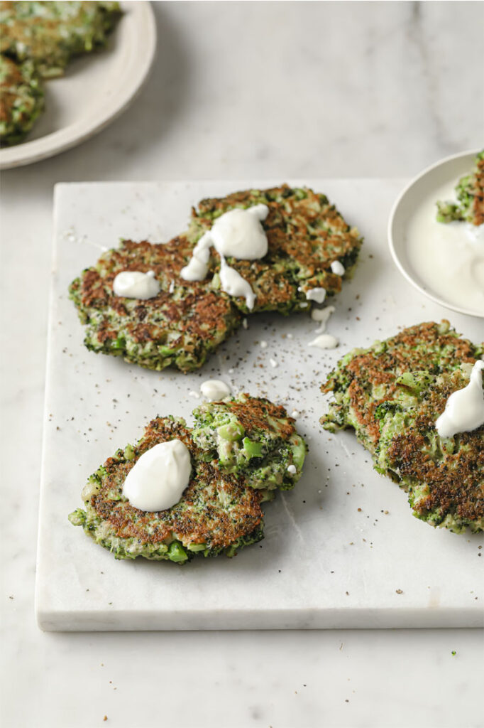 Angled, overhead shot of a marbled plate with golden brown broccoli fritters and sour cream dip. The broccoli fritters are garnished with sour cream, and in the background, you can see another plate with a few broccoli fritters. The plates are resting atop a marble countertop.