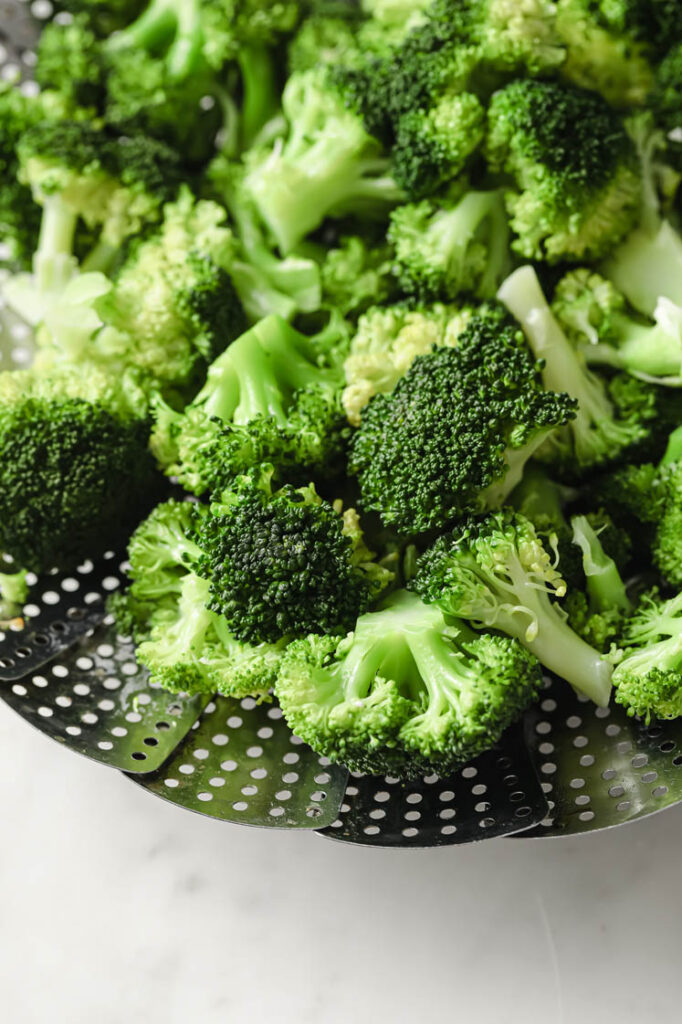 Close-up shot of fresh broccoli florets in a vegetable stainless steel steamer basket atop a marble countertop.