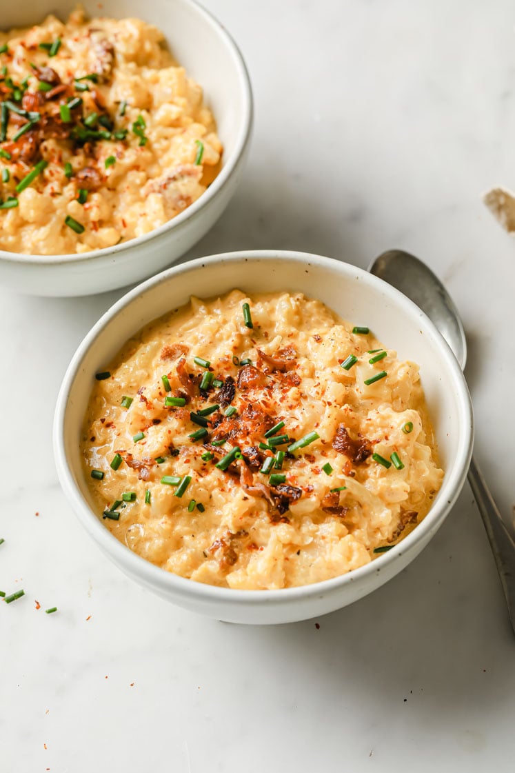 Overhead shot of a two small bowls with the cauliflower cheddar cheese bacon soup garnished with bacon crumbles, chives and red pepper flakes. In the background, there is a spoon. The bowls are sitting atop a marble countertop.