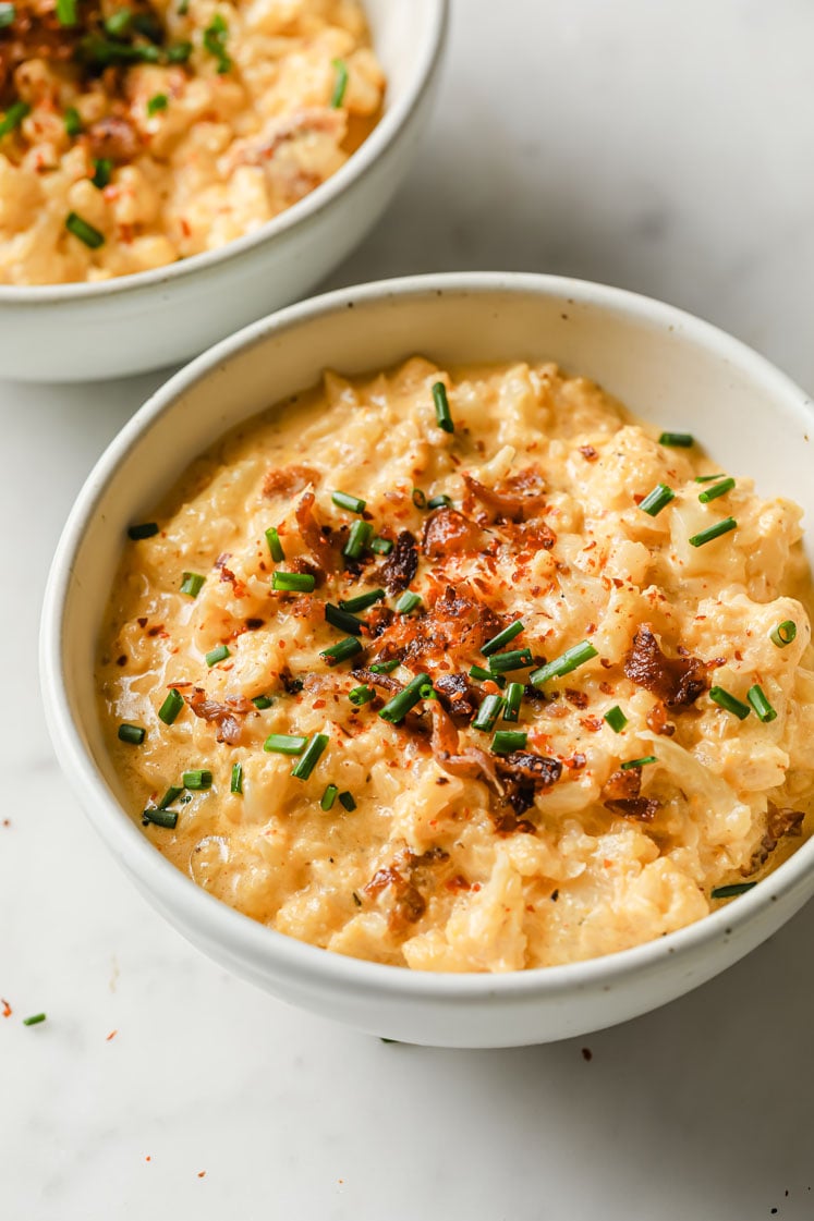 Close-up shot of small bowls with the cauliflower cheddar cheese bacon soup garnished with bacon crumbles, chives and red pepper flakes. The bowls are sitting atop a marble countertop.