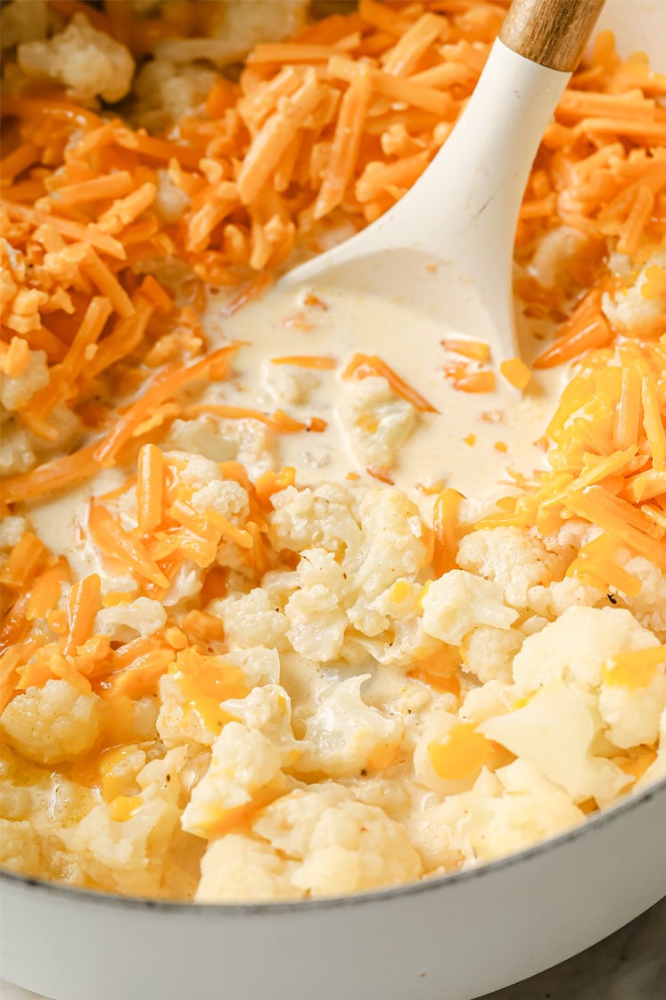 Overhead shot of a large pot with cauliflower florets, chicken broth, spices, heavy cream, butter, and shredded cheddar. Inside the pot, there is a ladle for stirring.